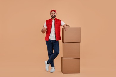 Photo of Happy young courier with stack of parcels showing thumb up on light brown background