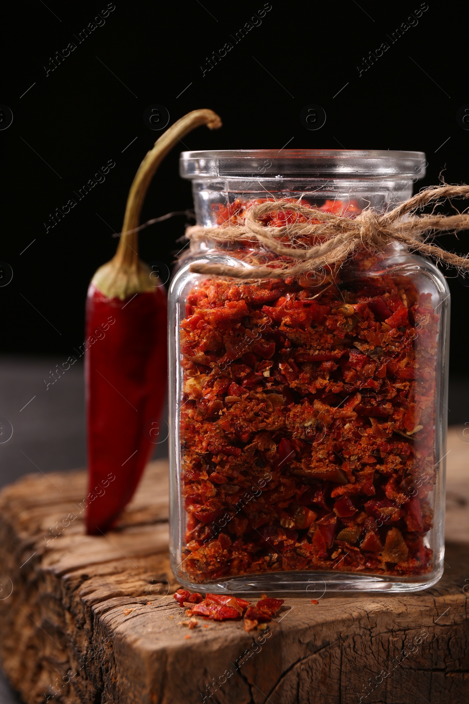 Photo of Chili pepper flakes in jar and pod on wooden board against black background, closeup