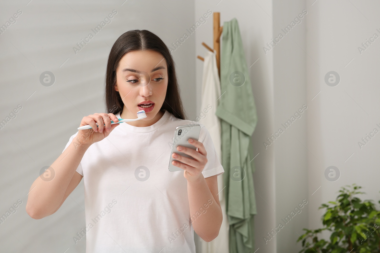 Photo of Beautiful young woman using smartphone while brushing teeth in bathroom. Internet addiction