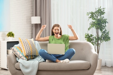 Photo of Emotional young woman with laptop celebrating victory on sofa at home