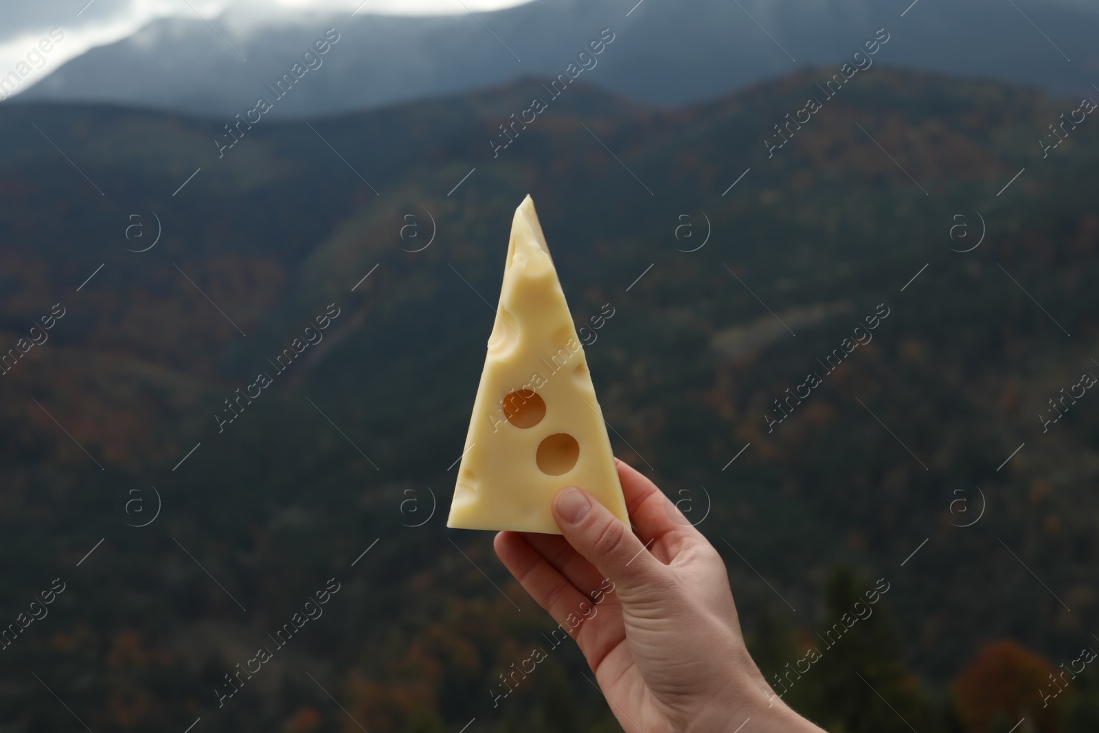 Photo of Woman holding piece of delicious cheese against mountain landscape, closeup