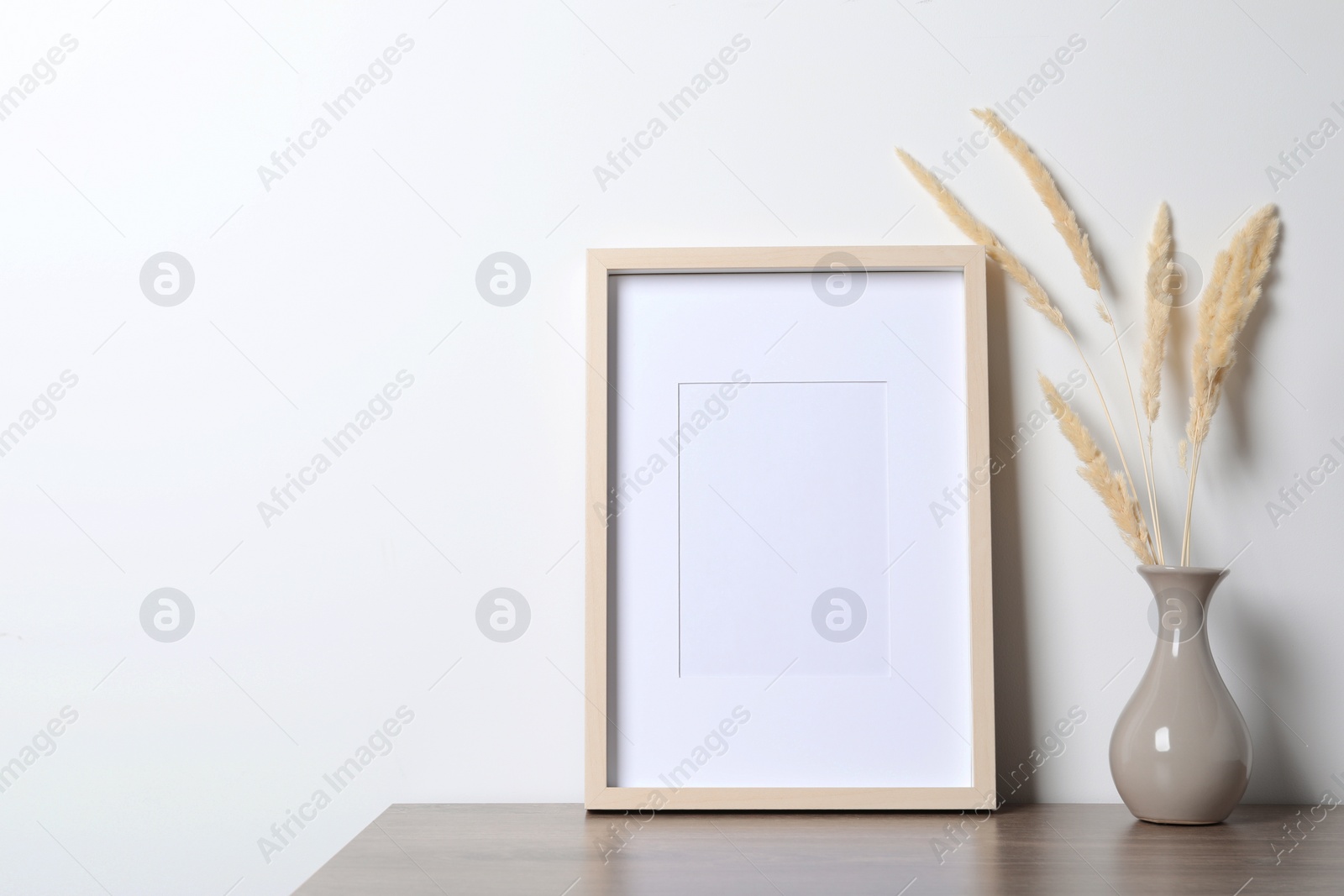 Photo of Empty photo frame and vase with dry decorative spikes on wooden table. Mockup for design