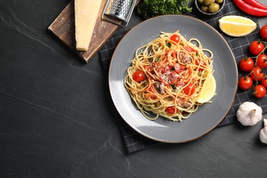 Plate of delicious pasta with anchovies, tomatoes and parmesan cheese near ingredients on black table, flat lay. Space for text