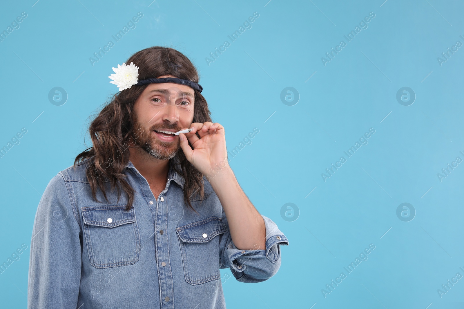 Photo of Stylish hippie man smoking cigarette on light blue background, space for text