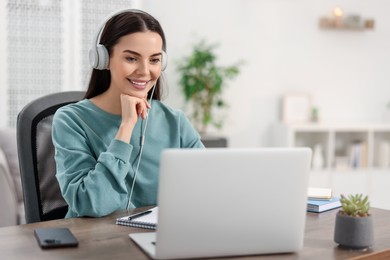 Photo of Young woman in headphones watching webinar at table in room