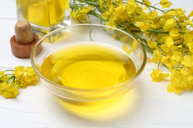 Rapeseed oil in bowl and beautiful yellow flowers on white wooden table, closeup