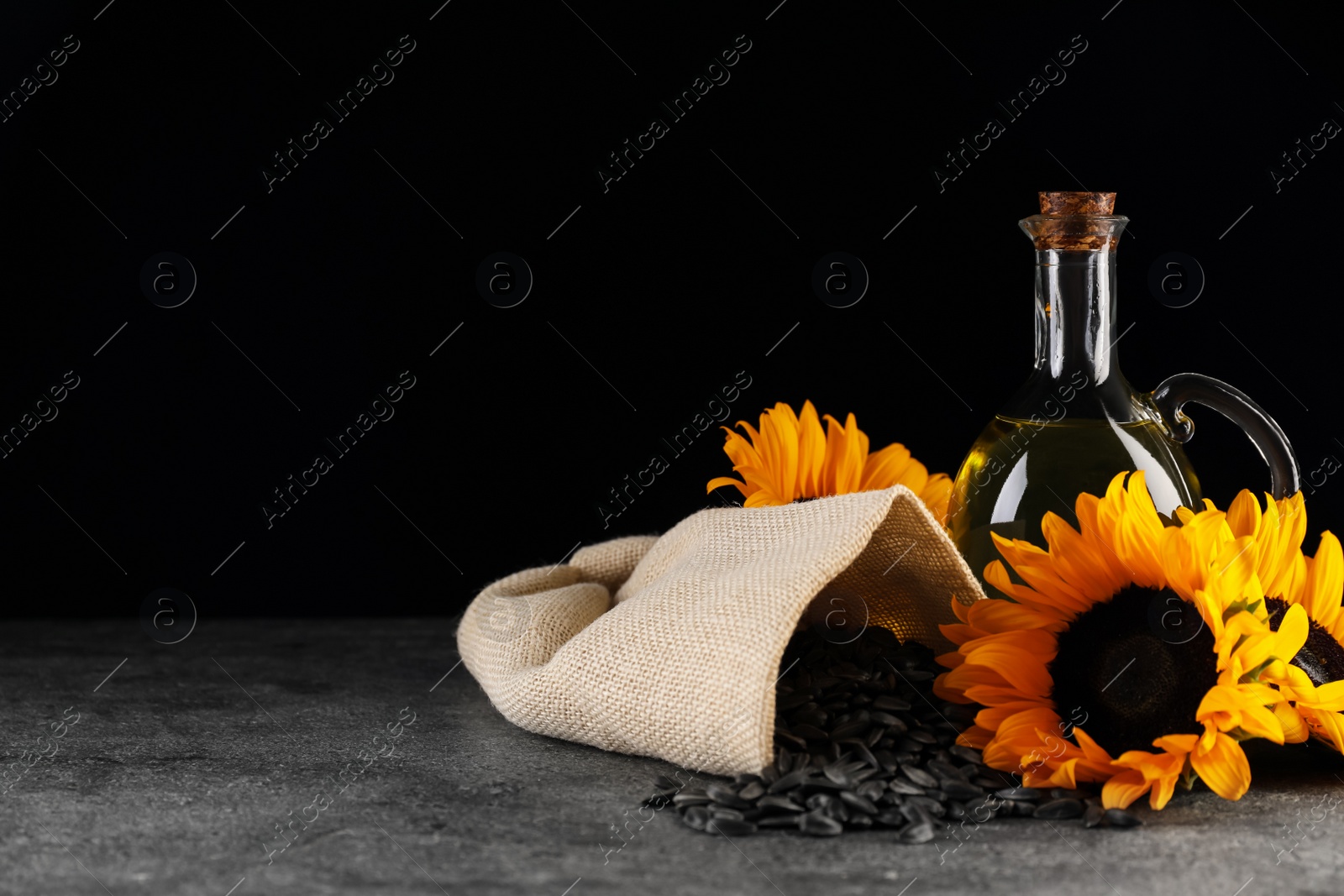 Photo of Sunflower cooking oil, seeds and beautiful flowers on grey table against black background. Space for text