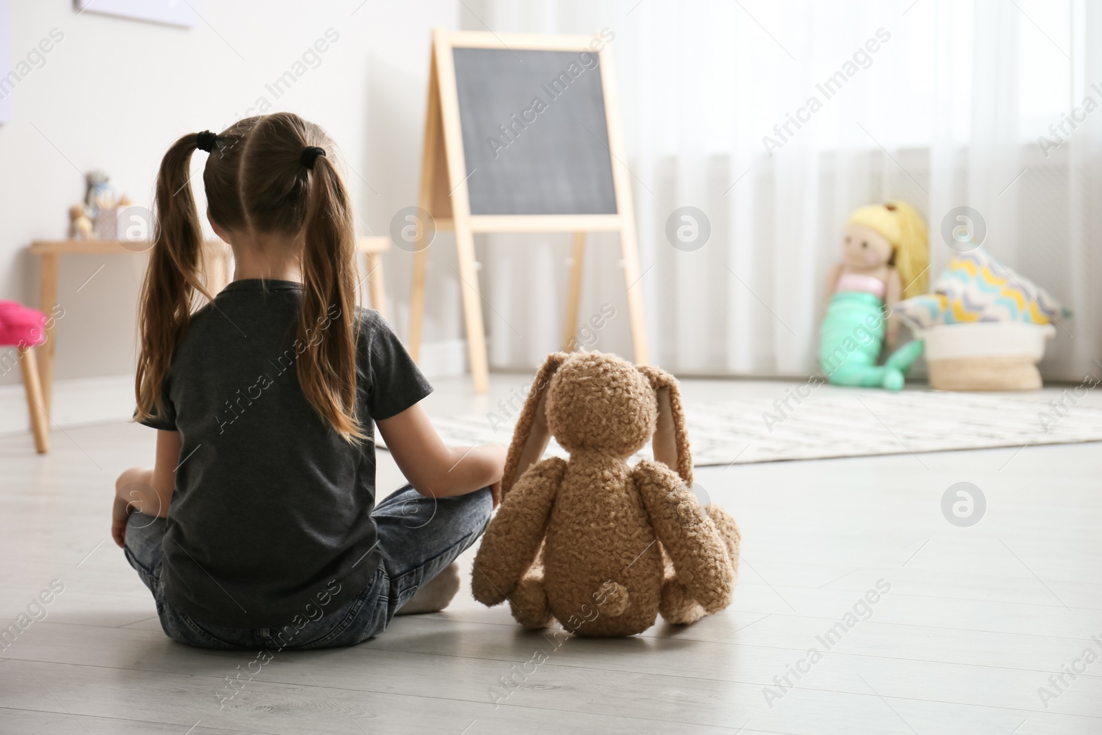 Photo of Lonely little girl with toy bunny sitting on floor at home, back view. Autism concept