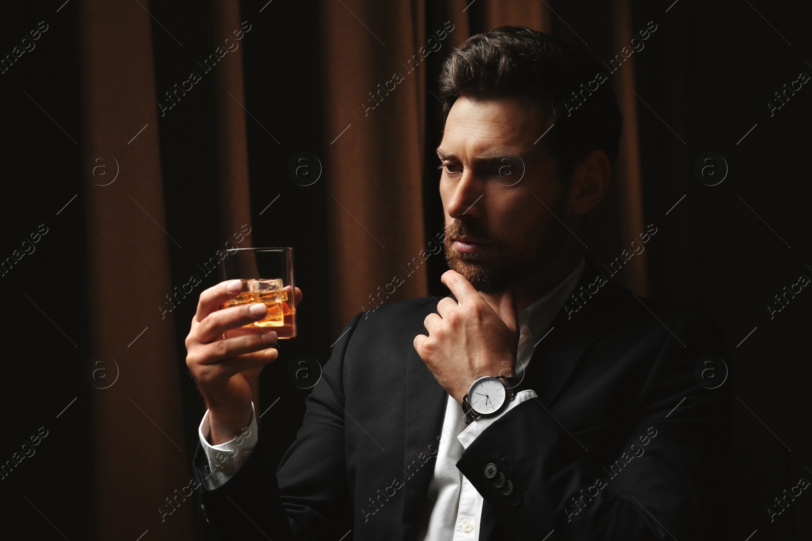 Photo of Man in suit holding glass of whiskey with ice cubes on brown background