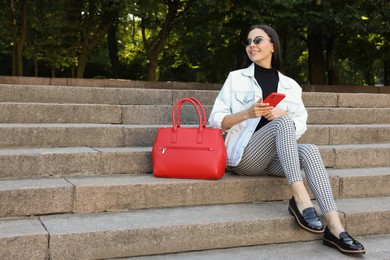 Photo of Young woman with stylish bag and smartphone sitting on stairs outdoors, space for text