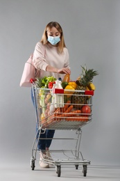Photo of Young woman in medical mask with shopping cart full of groceries on light grey background
