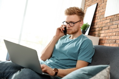 Photo of Young man talking on phone while using laptop in living room