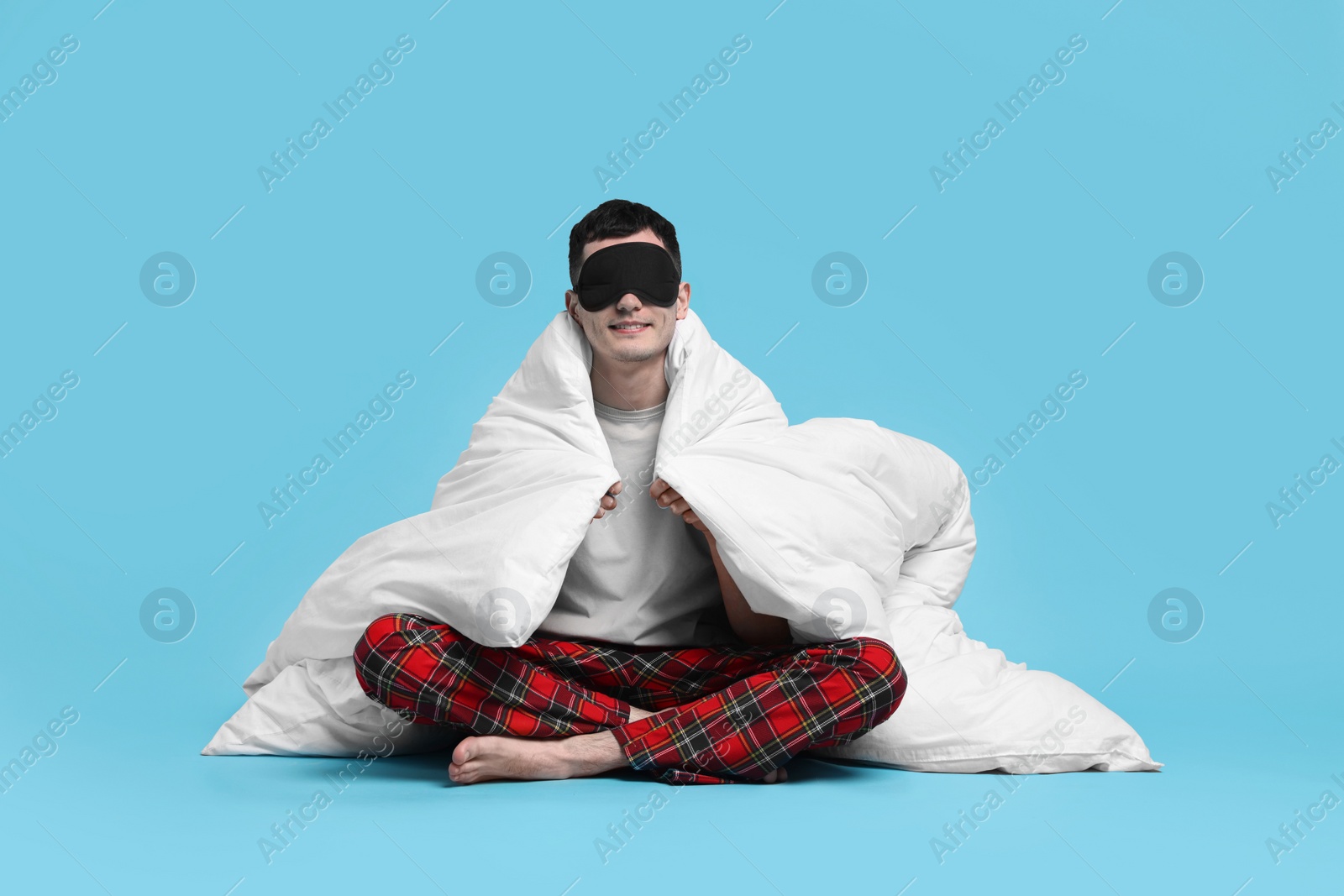 Photo of Happy man in pyjama and sleep mask wrapped in blanket on light blue background