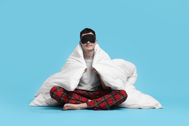 Happy man in pyjama and sleep mask wrapped in blanket on light blue background