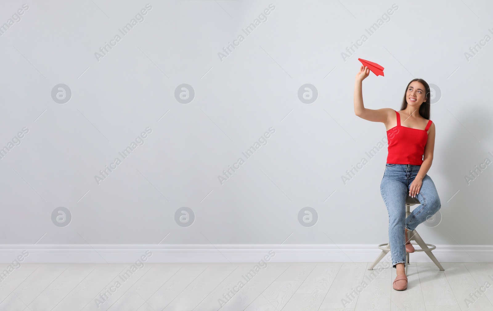 Photo of Beautiful young woman playing with paper plane near light grey wall. Space for text