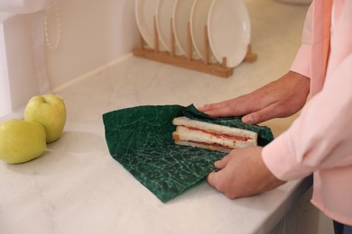 Man packing sandwich into beeswax food wrap at white countertop in kitchen, closeup
