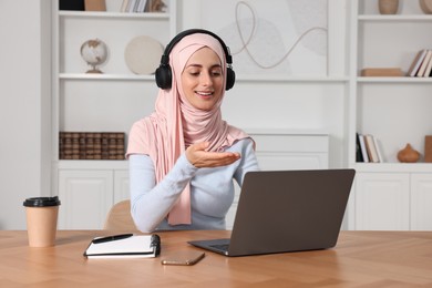Photo of Muslim woman in hijab using video chat on laptop at wooden table in room