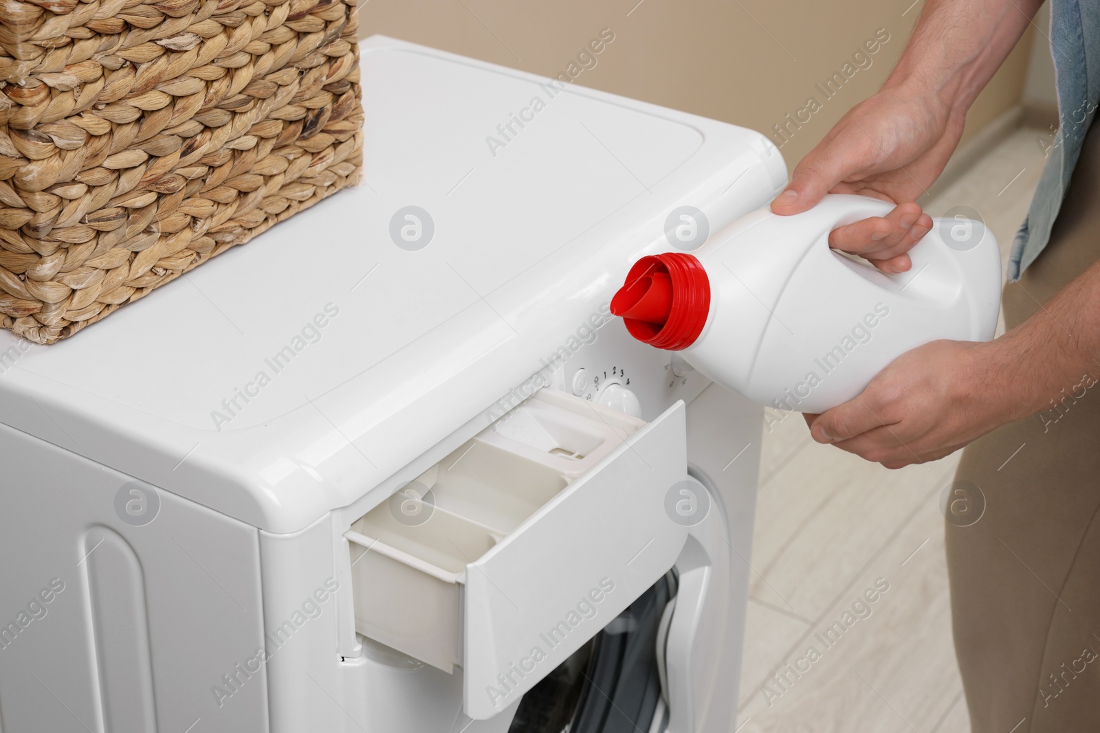 Photo of Man pouring fabric softener from bottle into washing machine indoors, closeup