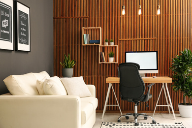 Comfortable workplace with computer near wooden wall in stylish room interior. Home office design