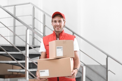 Photo of Young courier standing with parcels near stairs indoors