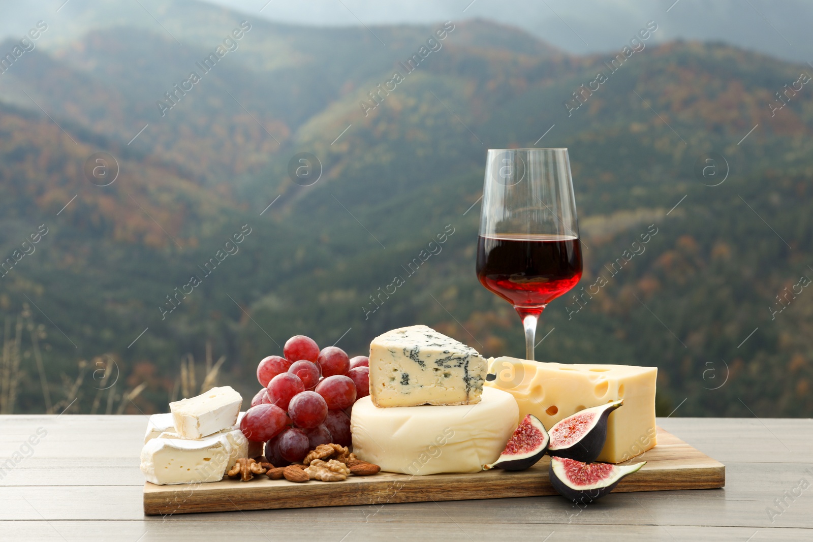 Photo of Different types of delicious cheeses, fruits and wine on wooden table against mountain landscape