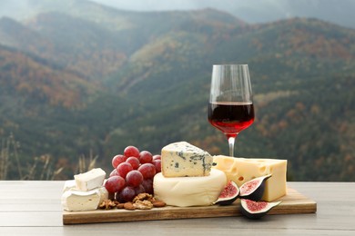 Photo of Different types of delicious cheeses, fruits and wine on wooden table against mountain landscape