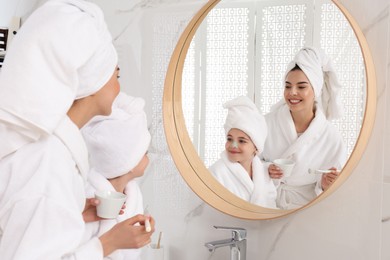 Photo of Young mother applying mask onto daughter's face near mirror in bathroom