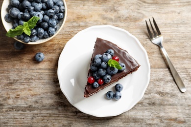 Plate with chocolate sponge berry cake and bowl of berries on wooden background, top view