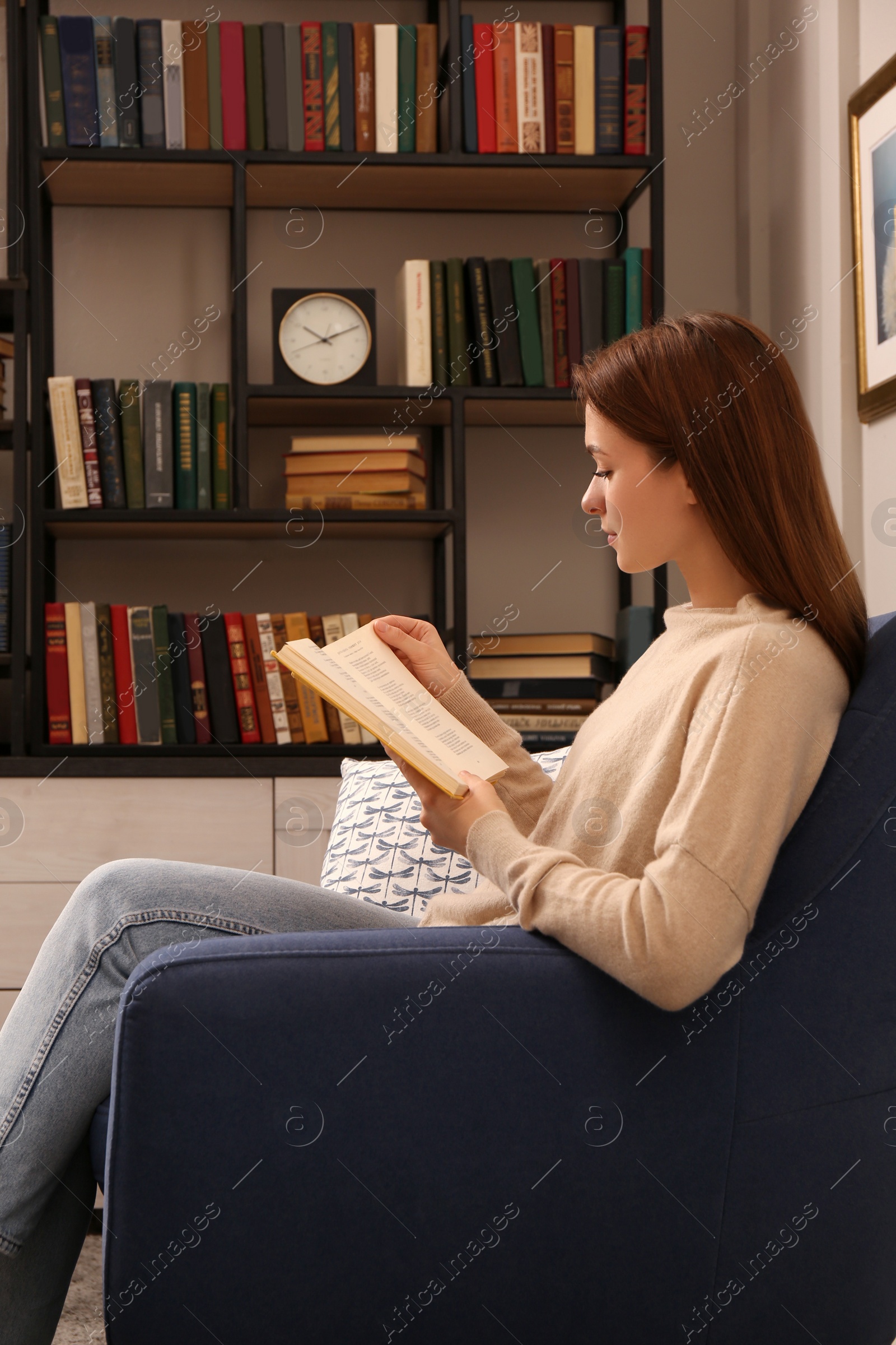 Photo of Young woman reading book in armchair indoors. Home library