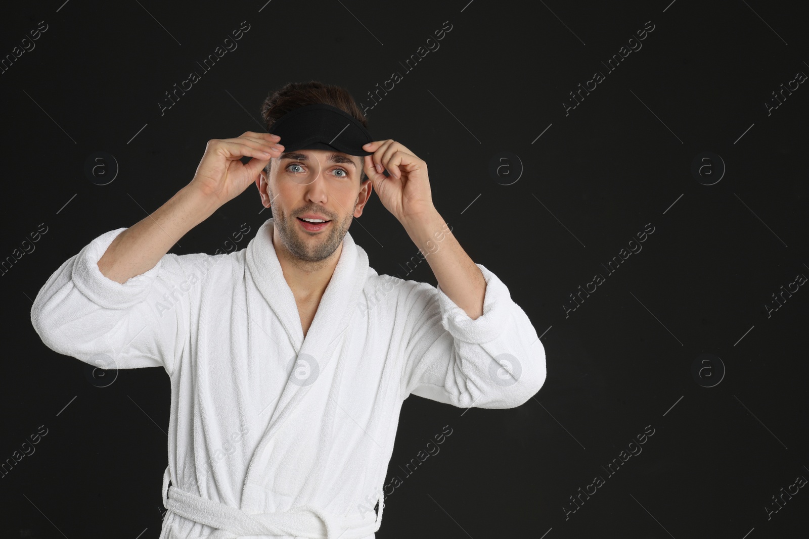 Photo of Surprised young man in bathrobe and eye sleeping mask on black background. Space for text