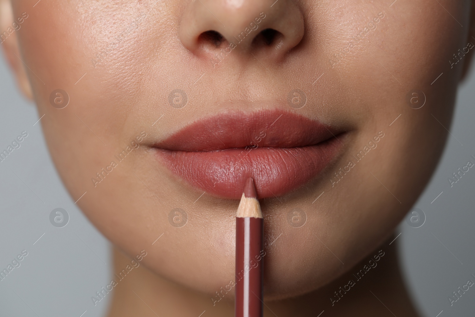 Photo of Young woman applying beautiful nude lip pencil on grey background, closeup