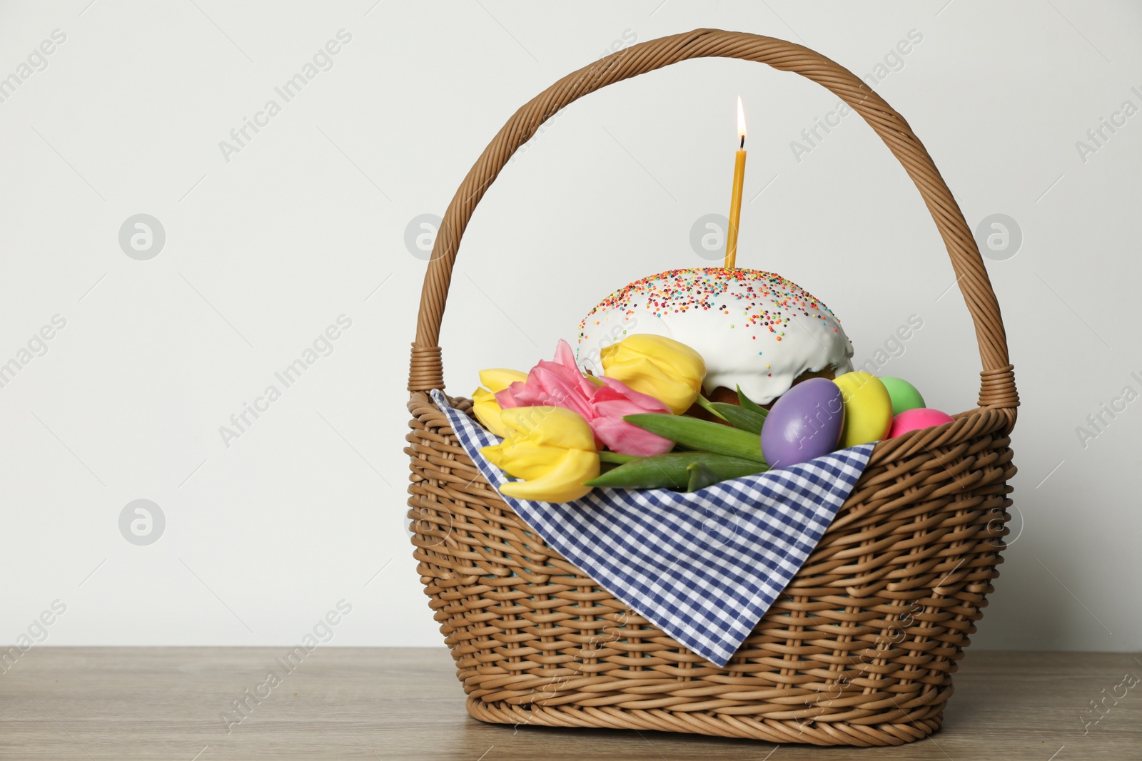 Photo of Traditional Easter cake with burning candle, dyed eggs and flowers in basket on table against white background. Space for text