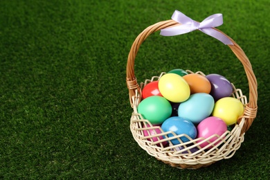Photo of Wicker basket with bow and bright painted Easter eggs on green grass. Space for text