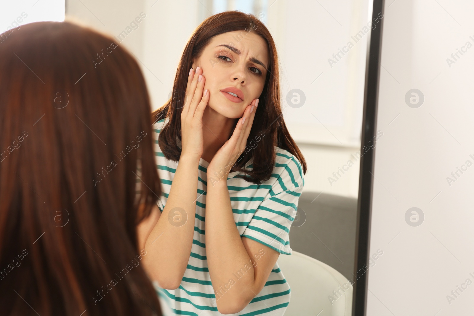 Photo of Young woman looking in mirror and touching her face indoors. Hormonal disorders