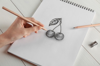 Photo of Woman drawing cherries with graphite pencil in sketchbook at white wooden table, closeup