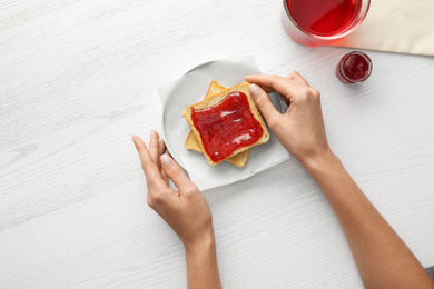 Woman eating toast with jam at white wooden table, top view. Delicious morning meal