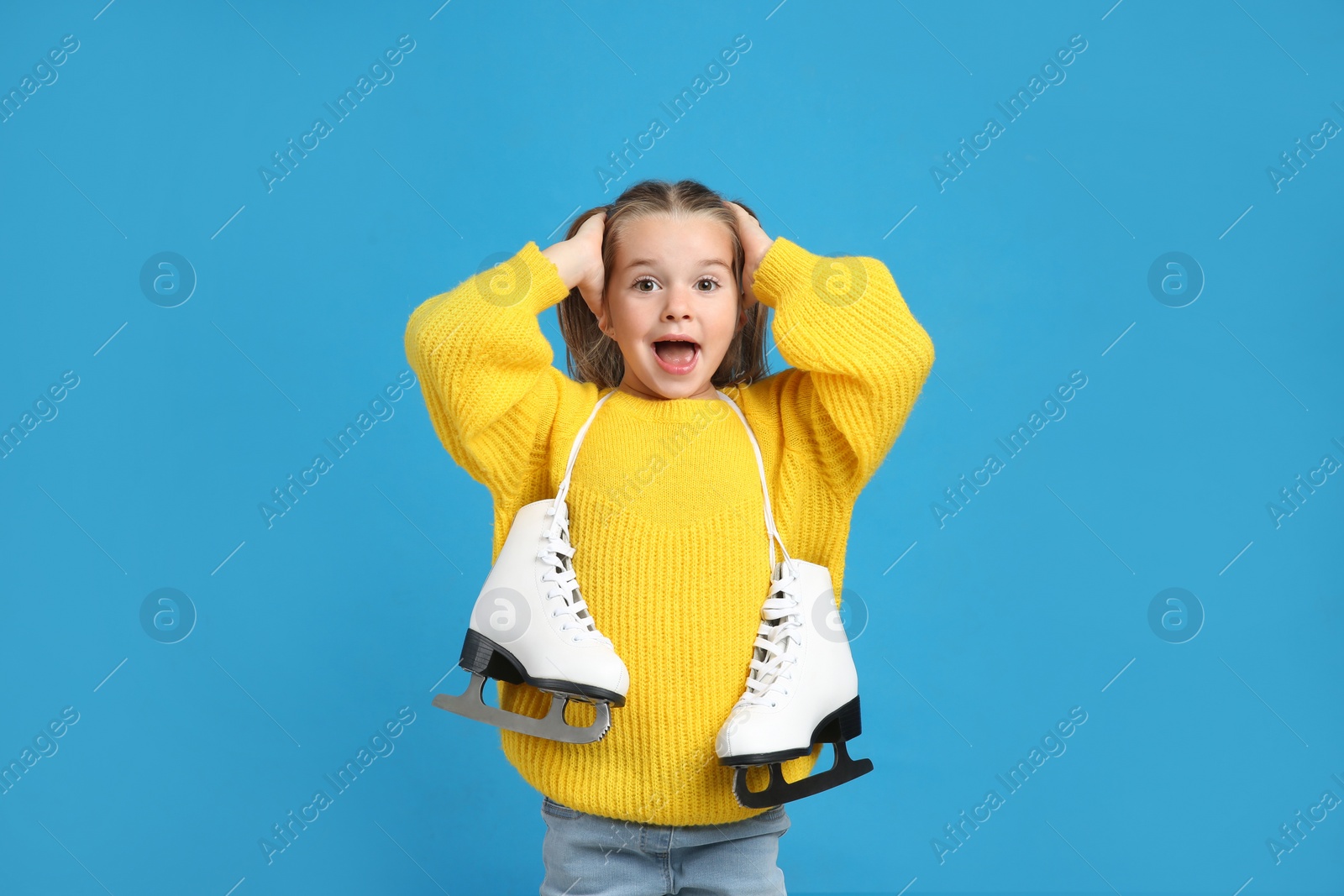 Photo of Excited little girl in yellow knitted sweater with ice skates on blue background