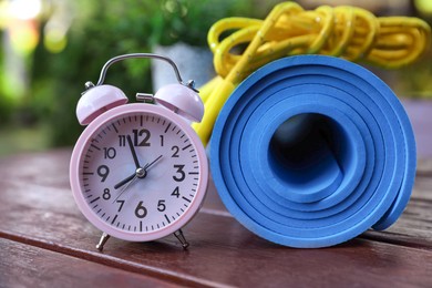 Photo of Alarm clock, fitness mat and skipping rope on wooden table outdoors, closeup. Morning exercise