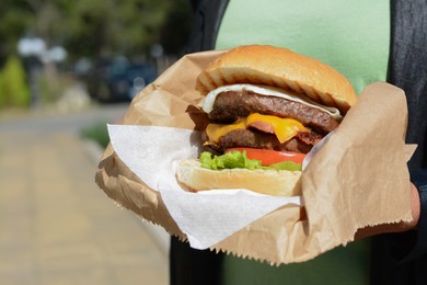 Woman holding delicious burger in paper wrap outdoors, closeup. Space for text