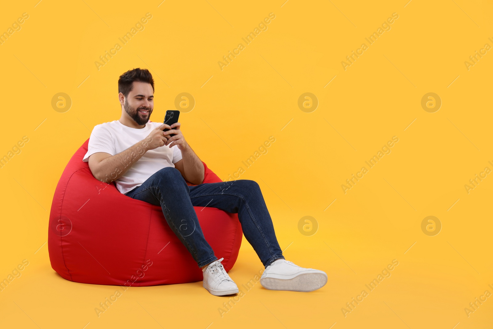 Photo of Happy young man using smartphone on bean bag chair against yellow background, space for text