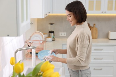 Happy young woman washing plate above sink in modern kitchen