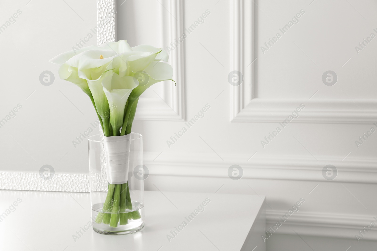 Photo of Beautiful calla lily flowers tied with ribbon in glass vase on white table, space for text