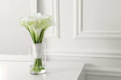 Beautiful calla lily flowers tied with ribbon in glass vase on white table, space for text