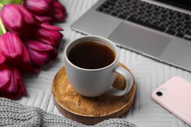 Beautiful tulips, cup of coffee, laptop, smartphone and sweater on light gray blanket, closeup