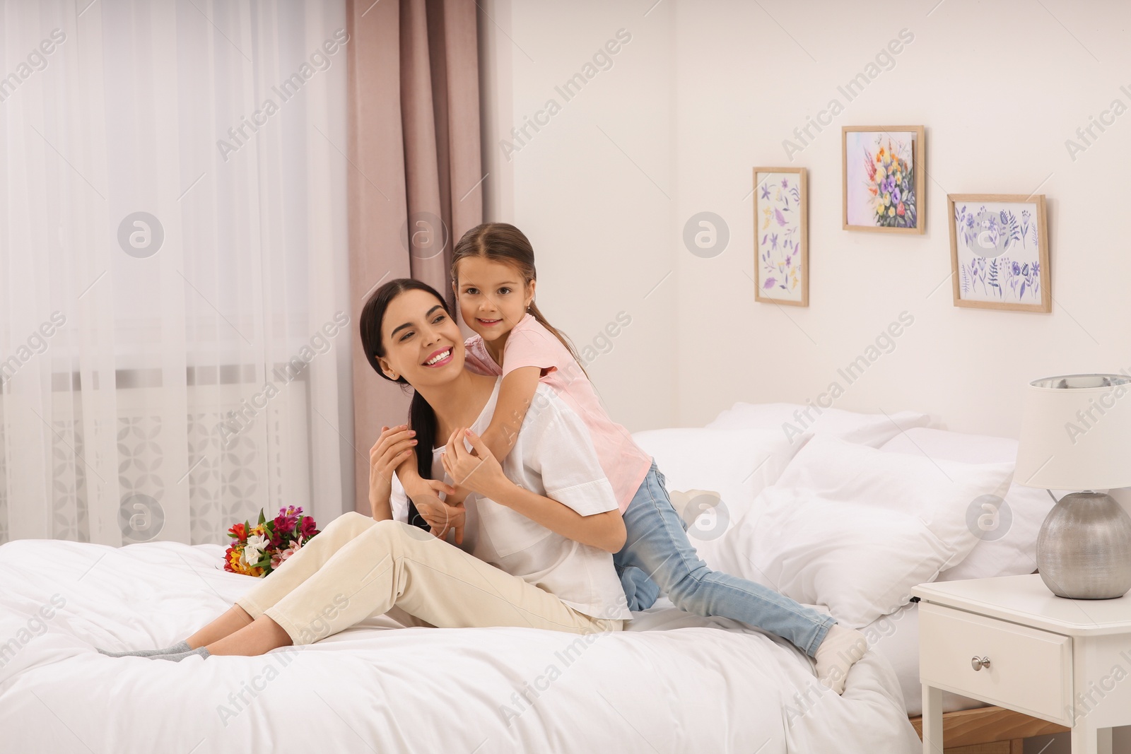 Photo of Happy woman with her daughter and bouquet of flowers on bed at home, space for text. Mother's day celebration