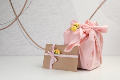 Photo of Furoshiki technique. Gift packed in pink fabric, card, and flowers on white table, space for text