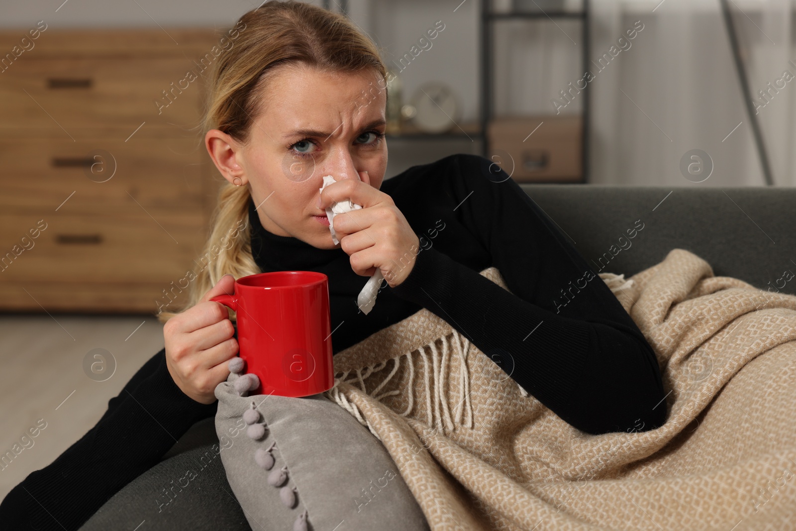 Photo of Sick woman with cup of drink blowing nose in tissue on sofa at home. Cold symptoms