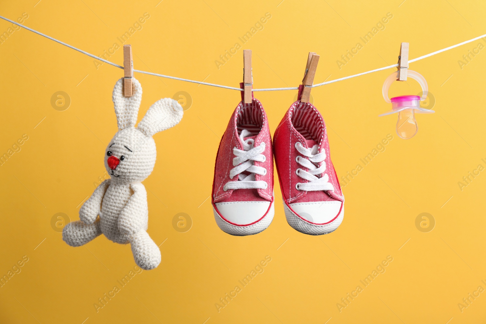 Photo of Small shoes, toy bunny and pacifier hanging on washing line against color background. Baby accessories