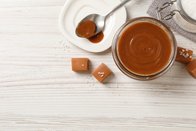 Photo of Yummy salted caramel in glass jar and candies on white wooden table, flat lay. Space for text
