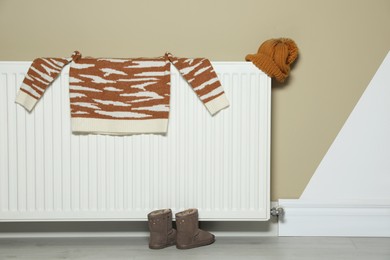 Photo of Heating radiator with knitted hat, sweater and shoes near color wall indoors